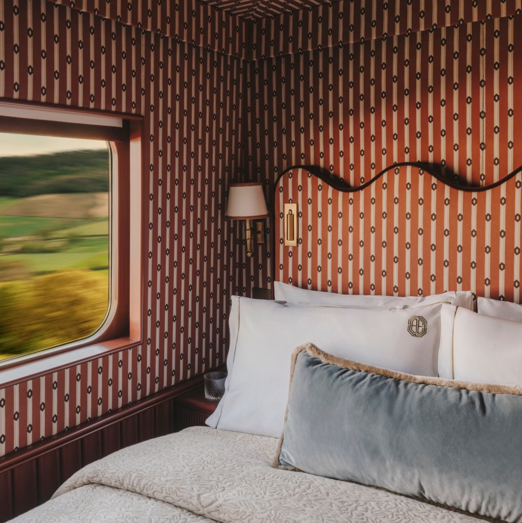 A cozy bedroom with patterned wallpaper, featuring a bed with decorative pillows and a window showing a scenic view of greenery outside.